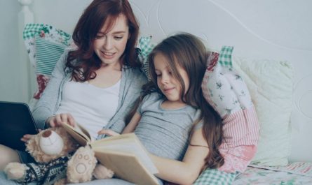 A mother and daughter enjoying a reading session on a cozy bed with a tablet and teddy bear.