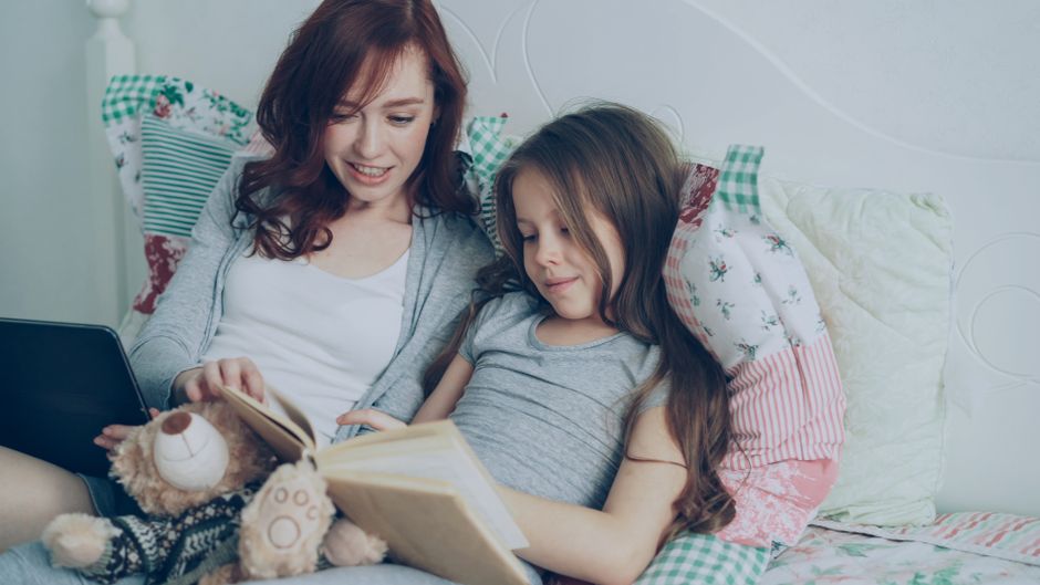 A mother and daughter enjoying a reading session on a cozy bed with a tablet and teddy bear.